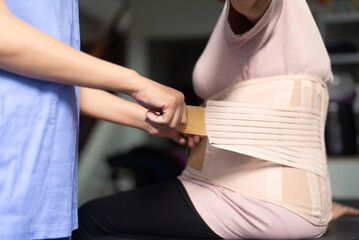 Physiotherapist helping senior women patient orthopedic corset at clinic. - obrazy, fototapety, plakaty