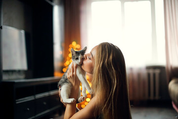 Pretty girl kiss kitten at Christmas tree at home in living room. Copy space and dark style. woman...