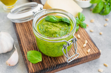 Traditional Italian basil pesto sauce in a glass jar on a board with ingredients for cooking on a gray concrete background. Copy space.