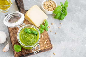 Traditional Italian basil pesto sauce in a glass jar on a board with ingredients for cooking on a gray concrete background. Copy space.