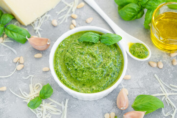 Traditional Italian basil pesto sauce in a white bowl with ingredients for cooking on a gray concrete background. Copy space.