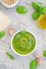 Traditional Italian basil pesto sauce in a white bowl with ingredients for cooking on a gray concrete background. Copy space.