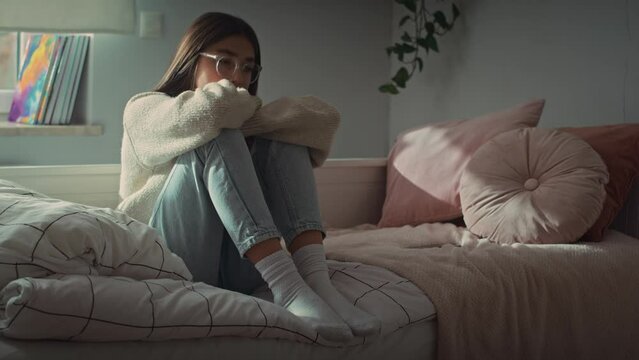 Pensive caucasian teenage girl sitting on bed with arms around knees and looking away. Shot with RED helium camera in 8K. 