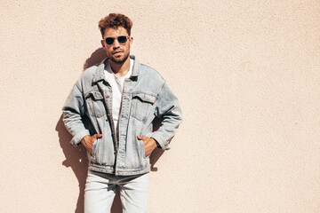 Portrait of handsome confident stylish hipster model. Sexy man dressed in jeans jacket. Fashion male posing in the street. In sunglasses in Europe city. Outdoors at sunset.  Near wall