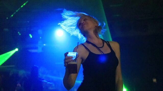 Sensual dancer woman drinking and dancing in a dark discotheque under blue-green laser strobe lights. Entertainment, leisure and nightlife concept. Adult lifestyle. Low angle view in slow motion.