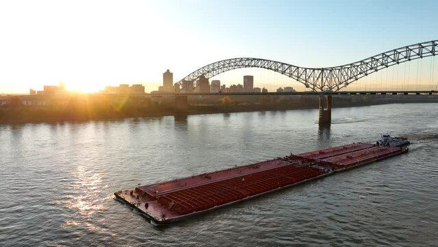 Barge on Mississippi River passes under Hernando de Soto Bridge in Memphis Tennessee USA. Aerial at sunrise.