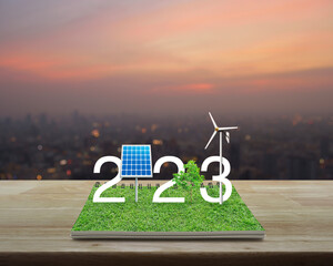 2023 white text with solar cell, wind turbine and growing tree on green grass on open book on...