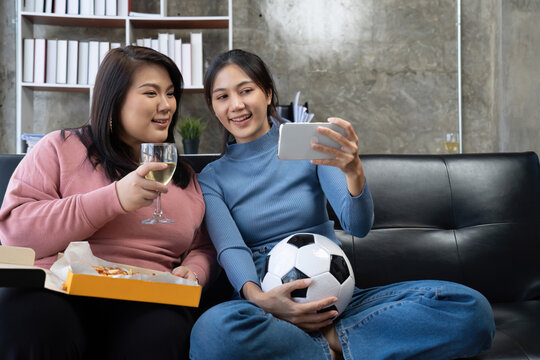 Friends watch sports on mobile phone, cheer and celebrate. Happy diverse asian friend supporters fans sit on couch with popcorn and drinks