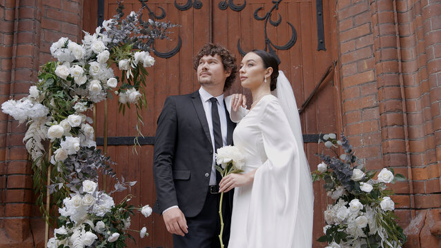 A bright and spectacular wedding couple. Action.Newlyweds where a bride in a white dress with a slit on her leg and a man with a beard in a suit walking next to a white arch with fresh flowers