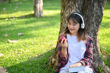 Asian female sitting under the tree, listening to music through her headphones and eating apple.