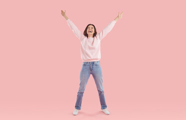 Young happy laughing brunette woman, dressed in a warm soft pink jumper and blue jeans, raises her hands palms up, closing her eyes with joy and laughter. Studio full-size photo on a pink background.