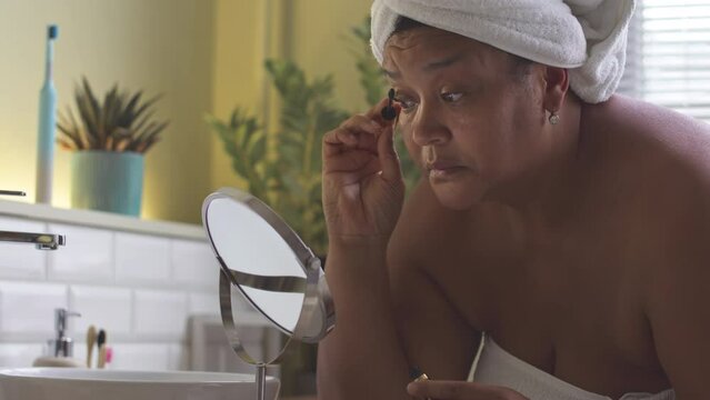 Mature African American woman in white towel looking at mirror while doing her makeup in morning, applying mascara