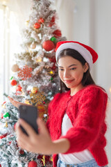 Cute beautiful young asian lady woman wearing santa hat selfie and video call with mobile phone posing in front a big Christmas tree with lots of decoration lights gift box and ornaments