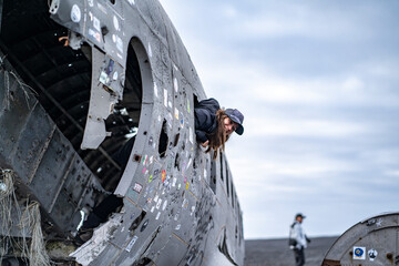 young woman peeking out of the window of damaged dc 3 plane in iceland black beach