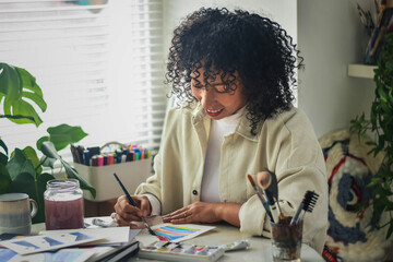 Smiling creative artist diverse bi racial woman with curly hair in white room home studio by window sitting at desk surrounded by art tools, plants and shelf, smiling and painting looking down