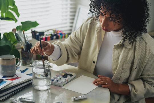 Creative artist diverse bi racial woman with curly hair in white room home studio by window sitting at desk surrounded by art tools, plants and shelf, cleaning paint brush, blank page