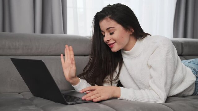 Brunette woman waves hello talking to boyfriend on video call via Notebook. Young female person lies on comfortable sofa using modern gadget