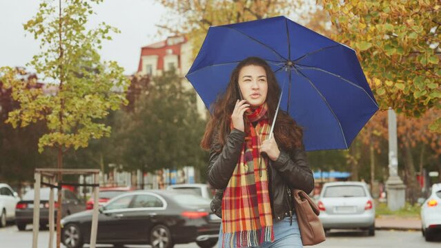 Smiling woman wearing leather jacket and checkered scarf talking on phone, blurred cars on background. Young female with blue umbrella under rain walking on autumn stree. Mobile addiction concept