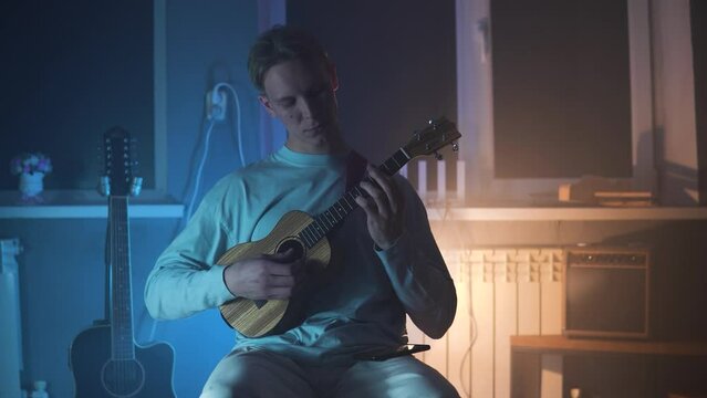 An adult smiling man plays the ukulele in the light neon dark room. A strong and blond guy sits and plays a small guitar in atmosphere place. A happy person plays a musical instrument.	