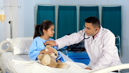 A young physician / pediatrician checking the heart rate of a cute little girl - a routine checkup. An adorable little kid wearing blue hospital clothes with an intravenous drip is sitting on a hos...