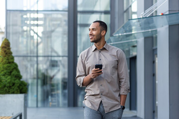 Smiling hispanic man walking down street near modern office building, freelancer businessman looking away holding mobile phone. - Powered by Adobe