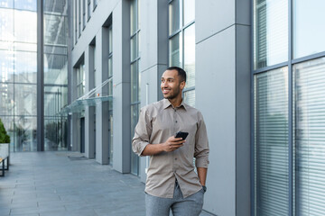 Smiling hispanic man walking down street near modern office building, freelancer businessman looking away holding mobile phone.