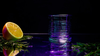 Close up of transparent glass standing on a bar counter with green decorative plant and orange fruit. Stock clip. Dark purple background at the night club.