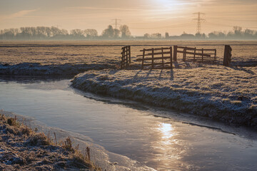 Dutch polder landscape on a cold day. The sun has just risen, it has frozen and there is ice on the ditch and frost on the grass. The photo was taken early in the morning in the Alblasserwaard region.