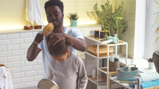 African American man brushing puffy hair of his little daughter standing by sink in bathroom in morning