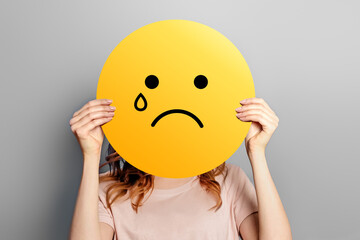 cry smile emoticon symbol. Girl holds a yellow poster with smiley with sad cry face isolated on a grey background - obrazy, fototapety, plakaty