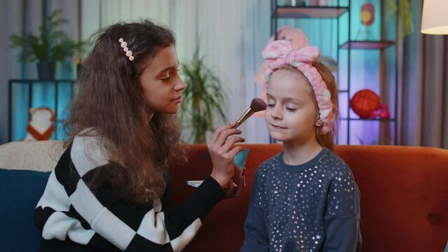 Teenage child helps or teaches to do face makeup her little sister kid girls. Female siblings children best friends putting applying make-up cosmetic at home play room. Friendship, family relationship