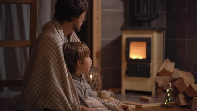A young man and his little son wrapped in a blanket sit by a domestic stove looking at a fire. Little cozzy wooden house. Winter vacations concept