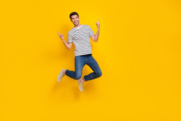 Fototapeta na wymiar Full length photo of cheerful cool guy dressed striped t-shirt showing hard rock signs jumping high isolated yellow color background