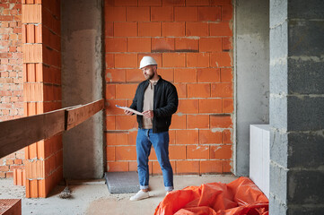 Full length of man worker in safety helmet holding papers and looking away while standing at construction site. Male builder contractor posing against brick wall in building under construction.