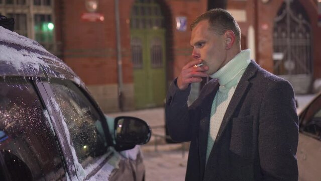 Man smoking cigarette and blowing smoke outdoors under snowfall near car parking at winter night. Concept of healthy lifestyle and bad habbits