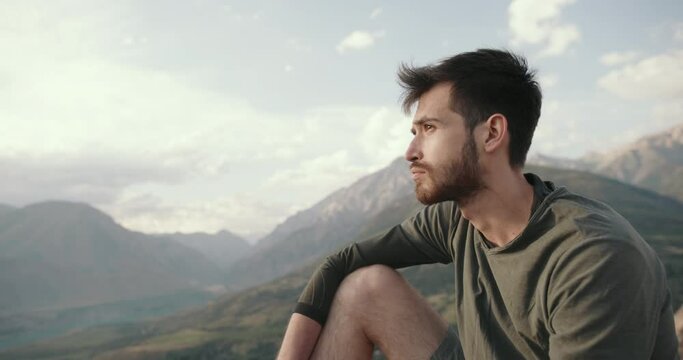 Portrait of an ethnic Caucasian man with a happy inspired bearded face. a young adult thoughtful guy is sitting on the top of a mountain against the background of the setting sun dreaming thinking