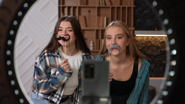 Two teenagers fooling around, dancing with fake mustaches. Young beautiful women posing for the camera using professional light, shooting a video for social media. High quality footage