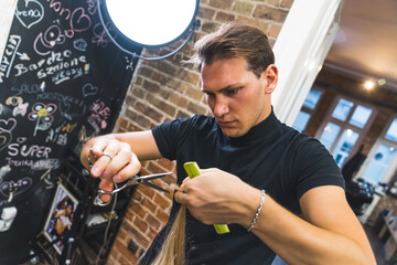 Professional focused male hairdresser styling his client's blonde hair with comb and scissors in the professional hair salon. Medium shot. High quality photo