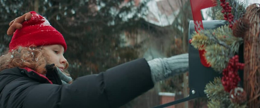 Portrait of cute little kid boy puts a letter with his Christmas wishes addressed to Santa Claus, Notrh Pole into the mailbox in the street