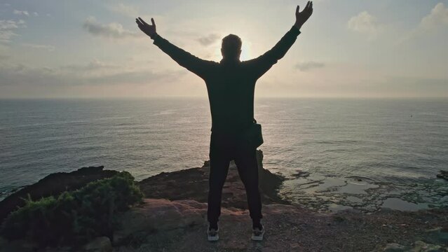 The camera follows a hipster millennial young man in a tracksuit running up a rock escarpment by the sea at sunset, throwing his hands up in the air, happy and drunk with life, youth and happiness