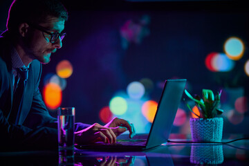 Serious young man, office manager and IT specialist sitting at office and using laptop at evening. Cool colored neon hue lights in office space.