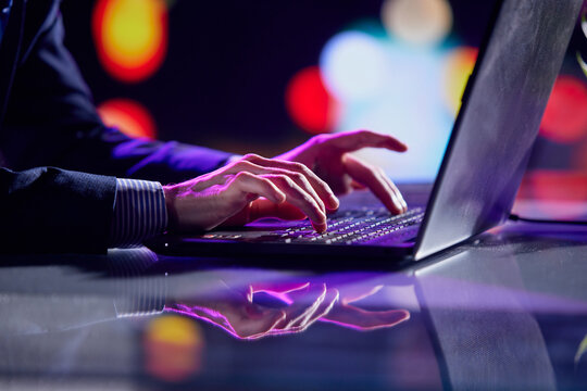 Cropped image of male hands of business man typing on laptop keyboard at evening time. Cool colored neon hue lights in office space.