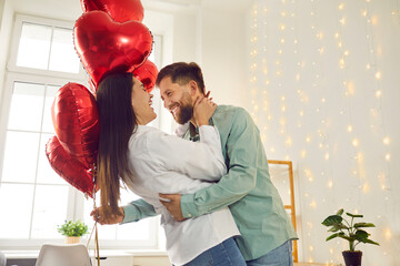 Fototapeta Happy, cheerful couple in love celebrating St Valentine's Day. Joyful, positive young man and woman enjoying Saint Valentine's Day, hugging, dancing with red heart shaped balloons, having fun together obraz