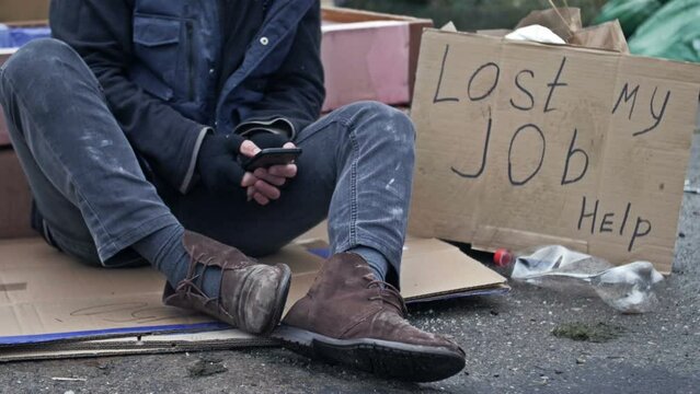 Homeless man shivering in the cold with a mobile phone in his hands sits on cardboard amongst the rubbish. Man appears to be looking through job advertisements. Next to him is a handwritten LOST MY