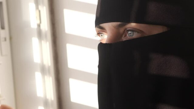 Woman with niqāb or niqaab opening window blinds. Light shining on Muslim lady. Refugee
girl with ruband locked up. No freedom Hijab    