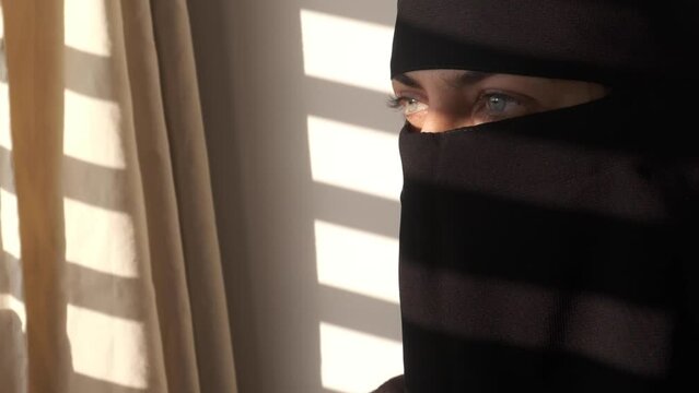 Muslim woman all covered in black looking out theMuslim woman opening window blinds. Light shining on covered face. Locked up girl. Hijab or Niqab
