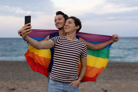 Beautiful gay young couple embraces and holds a rainbow flag. Happy couple taking selfie photo at the beach.