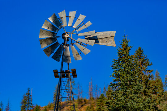 A small metal windpump stands against a bright blue sky and evergreen trees; Naramata, British Columbia, Canada