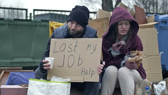 Two sad homeless men sit by the rubbish bins. Man and a woman are poor and dirty dressed. The man is holding a begging glass and a LOST MY JOB HELP poster. Young woman is sick and coughing.