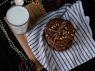 Chocolate cookies with pine nuts and glass of milk on white towel. Candles and golden beads on the black ground. Dark and moody morning food photography. - Powered by Adobe